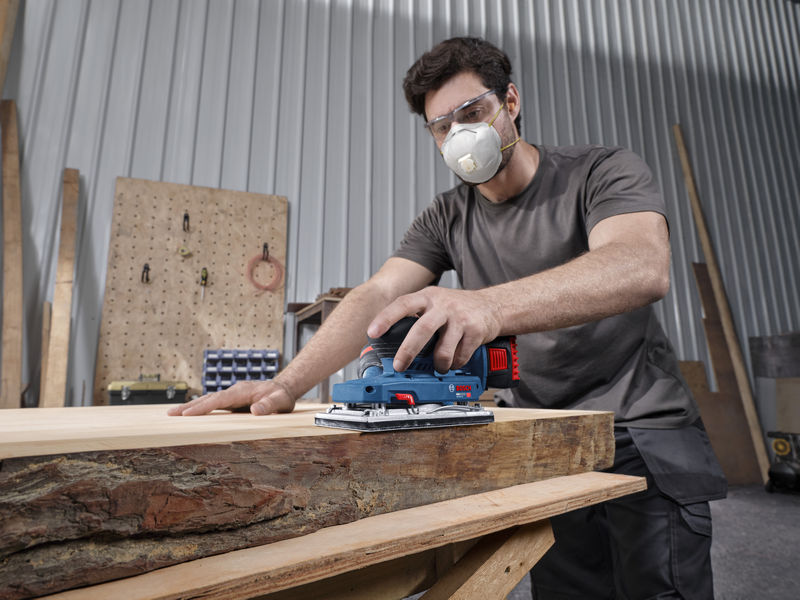 Anwendungsbild, handgehaltene Schleifmaschine, Holzbearbeitung, Staubmaske, Schutzbrille, Holzbrett